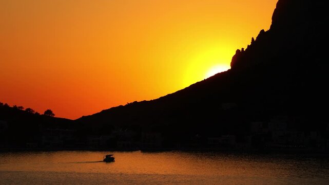 Breathtaking sunset over Telendos, a picturesque island in the Aegean Sea near Kalymnos. Warm golden and orange light bathes the rugged limestone cliffs, tranquil sea, and traditional waterfront