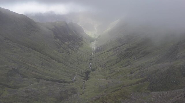 An aerial push-in shot captures a dramatic, misty valley below Stob Coire Sgreamhach, part of Bidean nam Bian and the Three Sisters in the Scottish Highlands, UK.