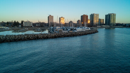 Obraz premium A beautiful high-angle drone photograph over calm blue water, capturing a massive stone breakwater pier protecting dry-docked winter boats against a backdrop of glowing suburban skyline condos.