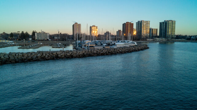 A beautiful high-angle drone photograph over calm blue water, capturing a massive stone breakwater pier protecting dry-docked winter boats against a backdrop of glowing suburban skyline condos.