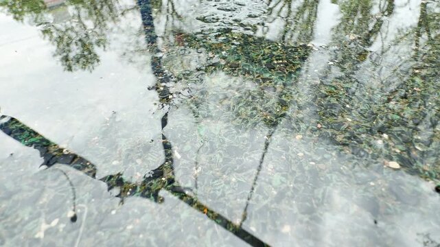 A wide rain puddle reflects the dark frame of a swinging swing set. The water shimmers over a textured playground floor during a quiet, gloomy afternoon outdoors.