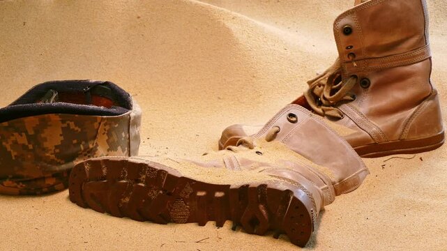 military helmet, a pair of worn combat boots, and a pair of broken sunglasses, partially buried in the sand. The arrangement suggests a narrative of someone having been in this harsh environment