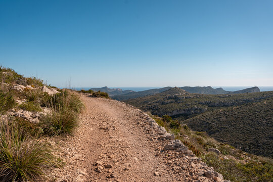 View of a winding dirt hiking trail through rocky hills and Mediterranean scrub with the sea in the distance Andratx, Balearic Islands, Spain.