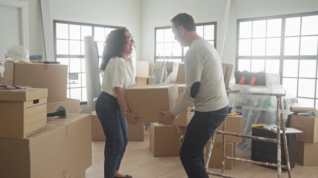 Man handing a cardboard box to woman while smiling, hands reaching among packed boxes and moving tools inside building; new home joy.