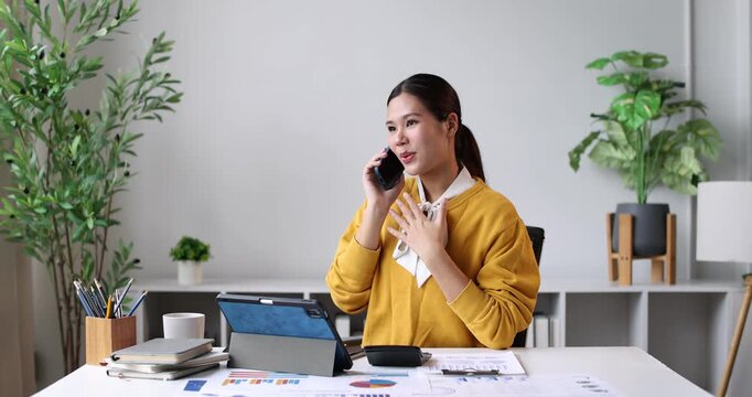 Asian businesswoman having a professional discussion on her smartphone, reviewing documents and analytics on a tablet, efficiently multitasking and working in a modern home office environment