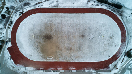 A striking top-down drone photograph looking straight down at an empty red running track oval surrounding a snow-covered soccer field and football uprights on a cold winter day. © Matt