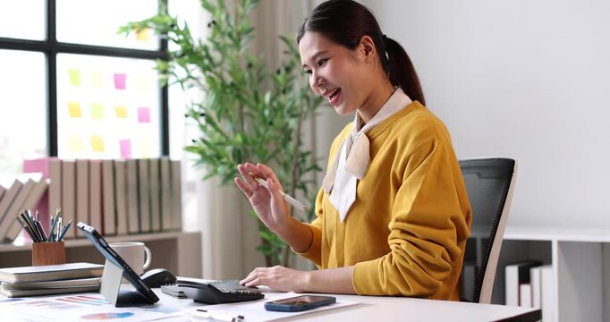 Asian businesswoman having a professional discussion on her smartphone, reviewing documents and analytics on a tablet, efficiently multitasking and working in a modern home office environment