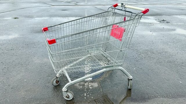 An empty steel shopping trolley rests stationary over a manhole cover on damp pavement. The expansive open area captures the distant, continuous low-frequency hum of city traffic.