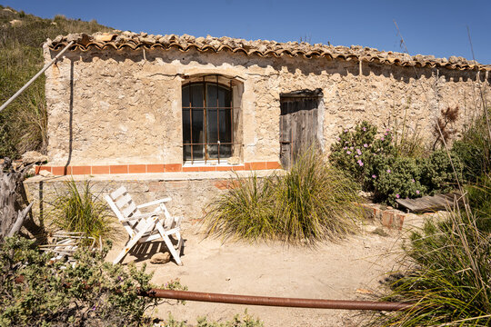 View of a rustic stone cottage with a tiled roof and a white wooden chair on a sunny day in Andratx, Balearic Islands, Spain.