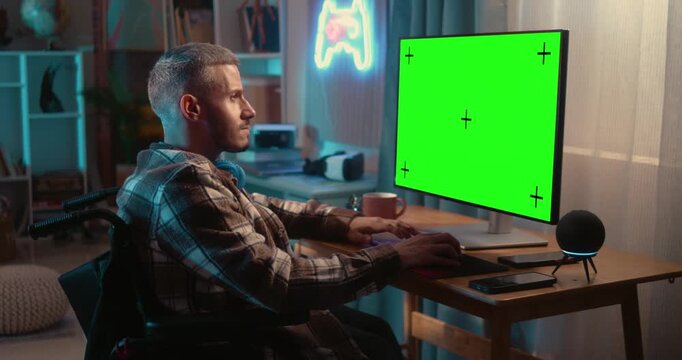 Focused man in wheelchair typing on a computer with a green screen monitor and reacting with excitement after achieving a successful result.
