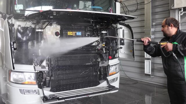 This image captures a professional cleaner utilizing a contactless washer to clean beneath the hood of a white truck in a large car wash hangar, showcasing effective vehicle maintenance techniques.