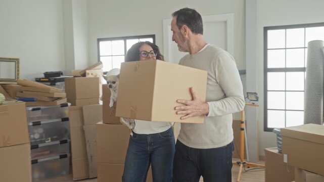 Couple carrying a cardboard box, woman peeks from behind it while man supports the load amid stacked packing boxes in a building during move; teamwork joy.