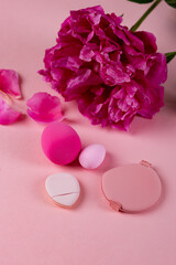 Fototapeta premium Cosmetic makeup sponges of different shapes, including beauty blenders, on a pink background with a pink peony and scattered petals. Beauty and makeup concept with skincare accessories.