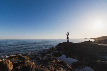 Fototapeta premium Silhouette of an 11 year old boy standing on a rocky coast in Crete and taking a picture of the sea with his smartphone