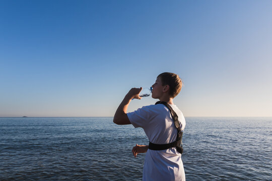 Boy with a broken arm wearing a medical shoulder sling brace drinks water from a bottle on the beach, focusing on hydration during summer recovery