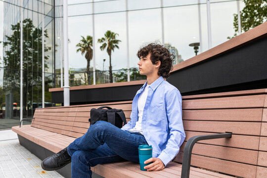 Curly haired man relaxing on bench with coffee in city