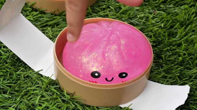 4K horizontal close-up video of a child's hands gently squishing a pink glitter dumpling squishy and taking it out of a clear plastic container. Satisfying sensory play with a soft sparkly fidget toy