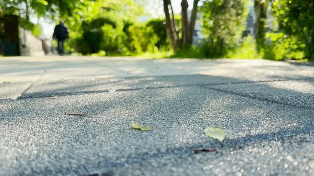 A blurred figure walks down a concrete paving stone sidewalk lined with green trees. Sunlight reflects off the textured ground in the foreground. The busy neighborhood echoes with the hum of the city.