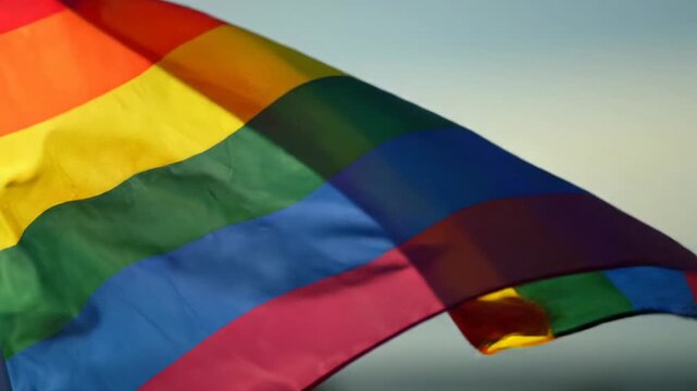 Vibrant rainbow pride flag in ECU, gently unfurling and rippling in soft golden hour light, showcasing sharp woven fabric texture and fluid motion, clean blurred background. Symbolizing pride and