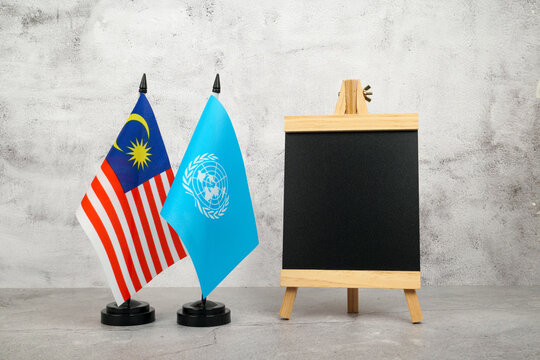 Malaysia and United Nations flags beside a blank chalkboard, symbolizing diplomacy, cooperation, football events, humanitarian efforts, and international collaboration.