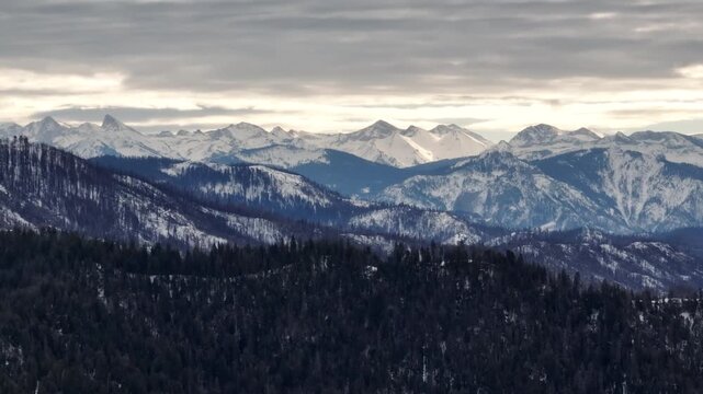 Aerial view of forested mountains with patches of winter snow under a cloudy sky in California