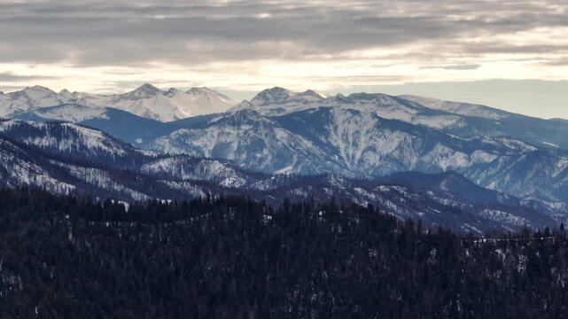 Aerial view of forested mountains with patches of winter snow under a cloudy sky in California