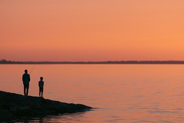 Fototapeta premium Father and son silhouette fishing together at sunset on a rocky shoreline, calm water reflecting warm colors of the evening sky
