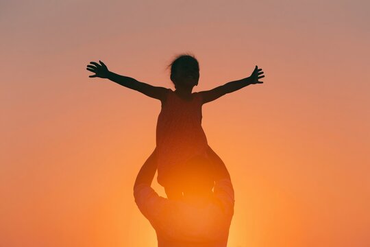 Father lifting daughter onto his shoulders against a vibrant sunset, silhouetted figures with arms outstretched, creating a joyful scene of connection and playfulness