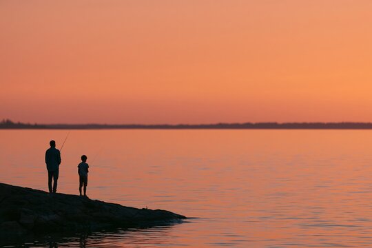 Father and son silhouette fishing together at sunset on a rocky shoreline, calm water reflecting warm colors of the evening sky