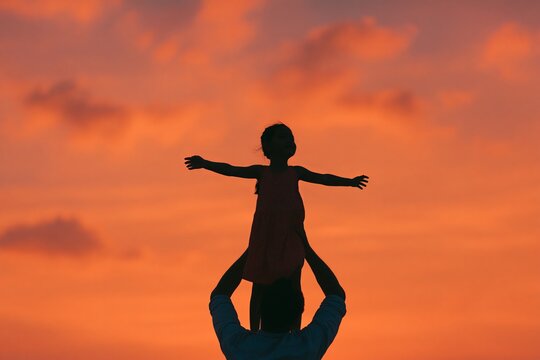 Father lifting daughter onto his shoulders against a vibrant sunset sky, silhouetted figures with arms outstretched, creating a joyful scene of connection and playfulness