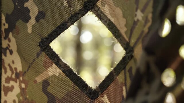 Camouflage Fabric Opening Blurred Nature Background - A detailed view of camouflage fabric with a diamond-shaped opening, revealing a softly blurred background of outdoor nature scenes with bokeh