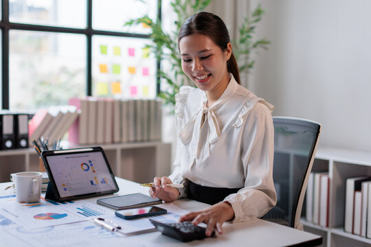Asian businesswoman analyzing financial data with a calculator and digital tablet, developing business strategies and managing finances in a modern office environment