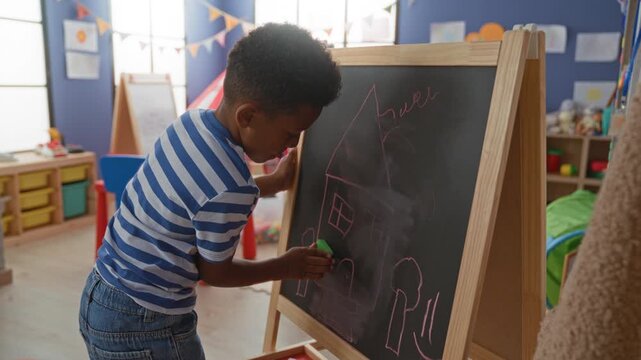 African american boy erases a chalkboard with hand while drawing a house on an easel in a daycare building filled with toys and bunting; playful focus.