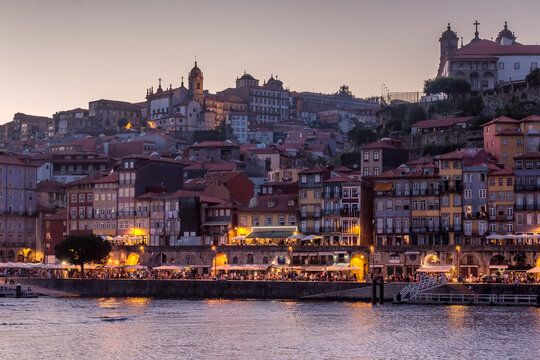 Porto, Portugal - 25 July 2016: View of the historic Ribeira district along the Douro River at dusk with illuminated buildings and people dining at waterfront cafes.