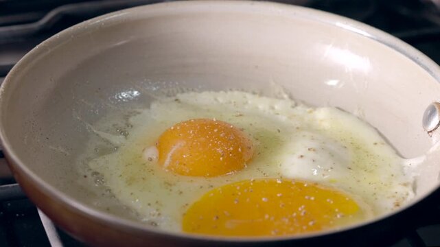 Frying and seasoning eggs in a pan. Yellow yolks glisten over a lightly cooked white.