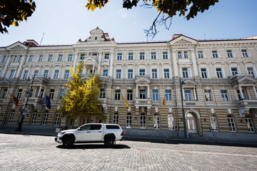 Naklejka premium Historic neoclassical Grand Hotel Vilnius, Curio Collection by Hilton building with white SUV on cobblestone street in Vilnius, Lithuania with flags and tree under a sunny sky