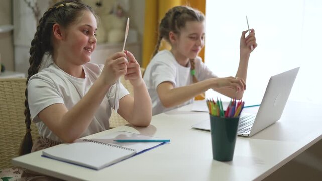 Two smiling girls learning online with laptop showing number cards at home. Happy students practice math together at table. Sisters study with computer, notebooks, and colorful pencils nearby.