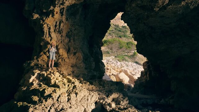 Woman backpacker walking through a rocky sea cave illuminated by sunlight, exploring hidden coastal landscapes and capturing travel memories during an outdoor adventure journey.