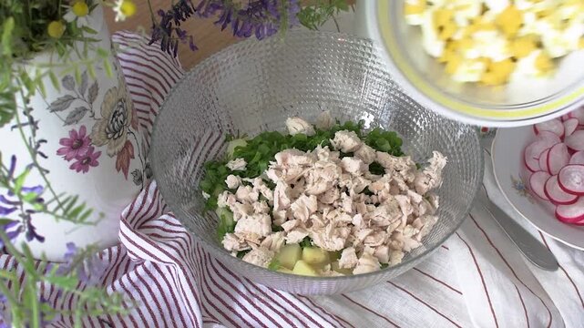 A woman prepares a cold summer salad called okroshka. She combines cooked potatoes, radishes, cucumbers, chopped chicken, fresh greens, eggs, and kefir in a bowl