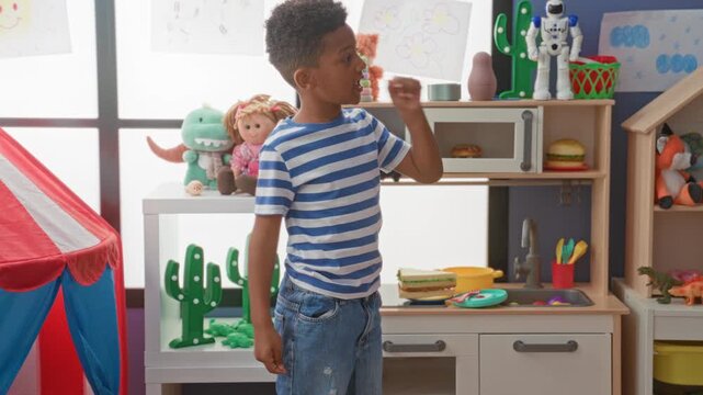 Black boy in striped shirt reaches hand to a colorful wall dartboard among stuffed toys in a playroom inside a building; childhood joy.