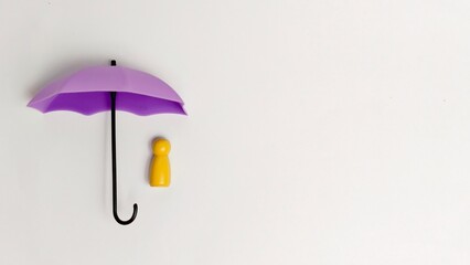 A miniature yellow peg doll stands beneath a small purple umbrella on a solid white surface, conceptualizing safety or insurance. © FaizAhmad