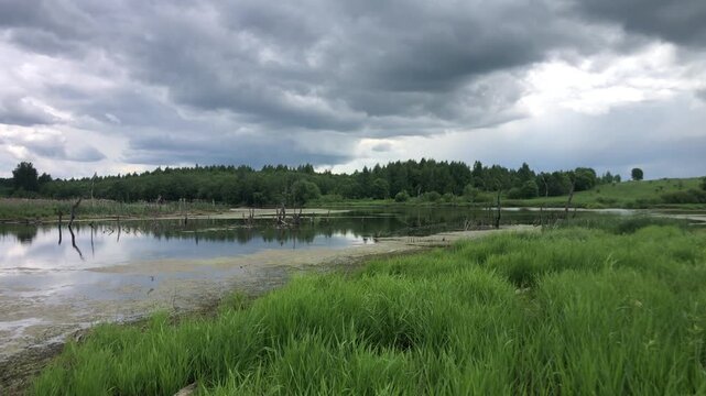 A landscape view of a calm river or lake under a dramatic, overcast sky filled with dark, heavy clouds. A line of dense green trees sits on the opposite bank, reflecting faintly in the water. The fore