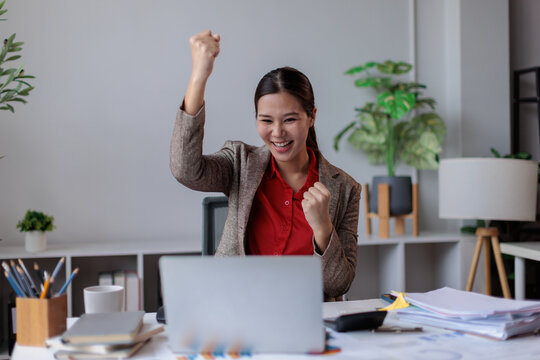 Asian businesswoman raising fists in a victorious gesture, smiling at a laptop screen, celebrating business goals and success in a modern office environment