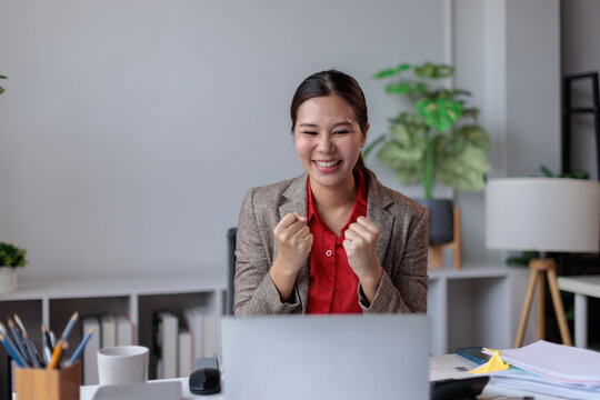 Happy young asian business woman celebrating work success or project completion. Clenching fists in excitement while looking at laptop screen in modern office
