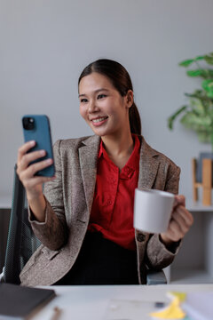 Businesswoman enjoying a coffee break while having a video call on a smartphone, connecting professionally with colleagues from her modern office workspace