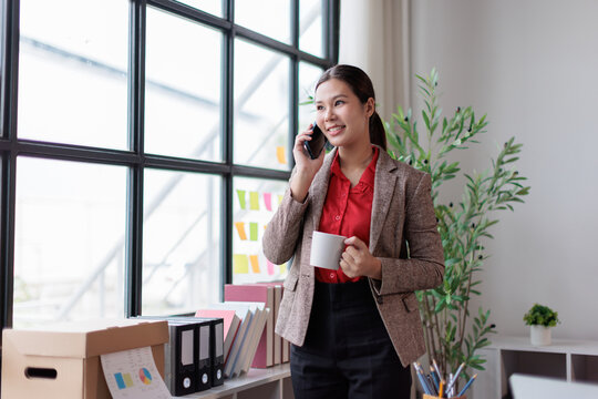Asian businesswoman having a phone conversation in a bright office environment. Expressing happiness and confidence while holding a coffee mug. Representing modern work communication