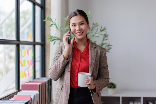 Asian business woman smiling, using a mobile phone for communication while holding a coffee cup, standing near an office window during her busy workday