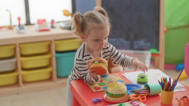 Little blonde girl places toy burgers and a plate on a red table near colored pencils and a chalkboard in a preschool building; concentration.