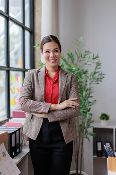 Asian business woman smiling with arms crossed, standing in a smart office environment, portraying confidence, leadership, success, professionalism, and management in her role