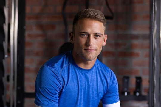 Man in late twenties wearing blue tee with compression sleeve sitting in gym by weight rack